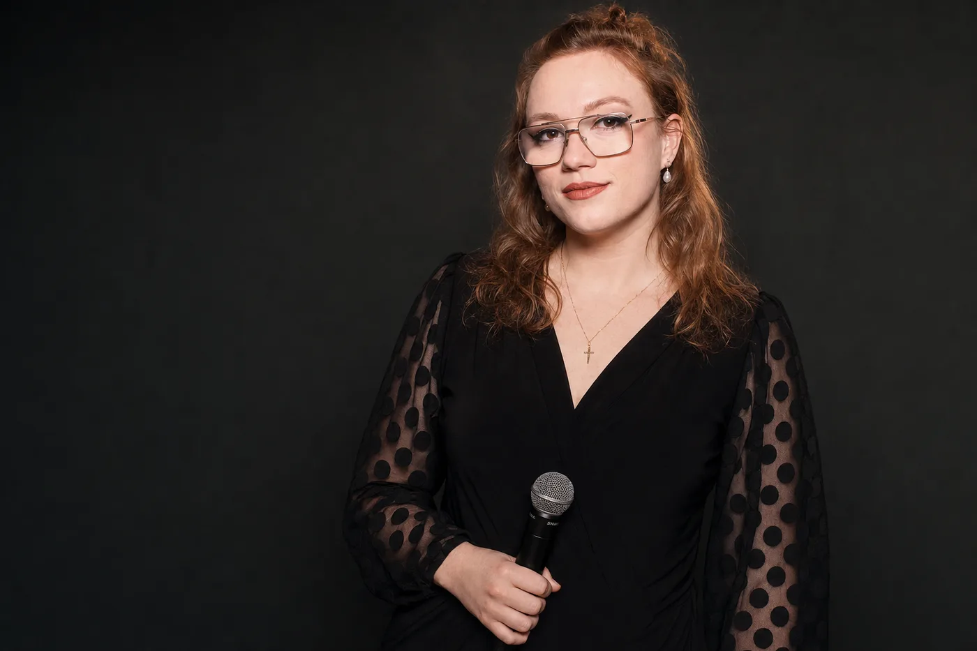 MC/DJ Allie of Sound of Harmony holding a microphone in a black polka-dot dress against a dark studio backdrop, professional wedding MC, DJ and stand-up comedian serving Niagara and Southern Ontario